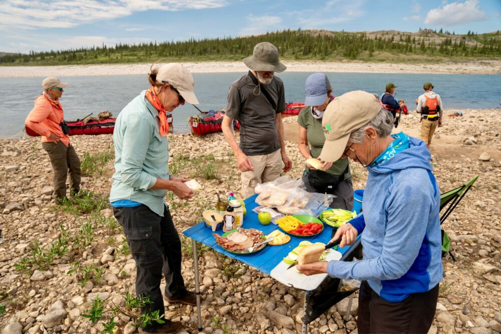 Guests serving themselves from a deli lunch spread on the Horton River shore, with beached canoes in the backdrop.
