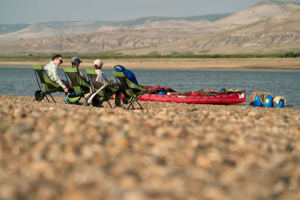 Guests relaxing in camp chairs on Horton River's shore, with mountain views.