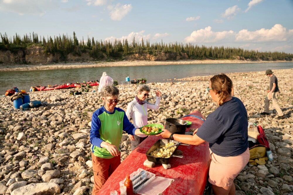 Guests having dinner at a riverside campsite, using an overturned canoe as a makeshift table on the Horton River.